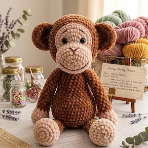 May include: A handmade, plush crochet monkey in shades of brown and tan. The monkey has a smiling face, large ears, and is seated on a light-colored surface. A small sign with handwritten text is visible, along with jars and spools of yarn.