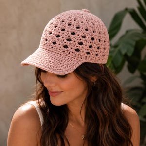 May include: A light pink crocheted baseball cap with a patterned crown and a solid brim. The cap is made of a soft, textured yarn and has a small button on top. The hat is designed for sun protection and casual wear.