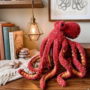 May include: A large, handmade red crochet octopus with yellow accents on its tentacles. The octopus is sitting on a wooden surface, with books and decorative items in the background. The octopus is a unique, handcrafted item.