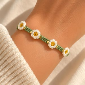 May include: A green macrame bracelet adorned with white daisy flowers with yellow centers and small gold beads. The bracelet is worn on a wrist, showcasing the delicate floral design and craftsmanship.