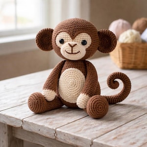 Puede incluir: Un mono de ganchillo hecho a mano en tonos marrón y crema. El peluche presenta una cabeza redonda con orejas grandes, una cara color crema y ojos negros. El mono está sentado con los brazos y las piernas extendidos, y su cola está enroscada.