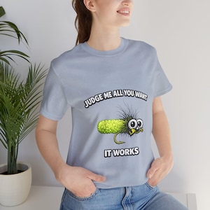 May include: Light blue t-shirt with the text "JUDGE ME ALL YOU WANT IT WORKS" above and below a cartoon pickle with a fishing hook. The pickle has googly eyes and black hair.