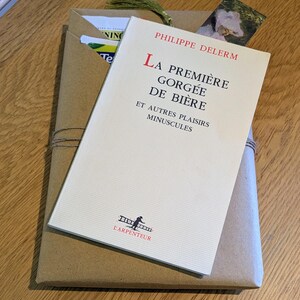 May include: A book titled "La Première Gorgée de Bière" by Philippe Delerm, with a brown paper wrapping and a green tassel. The book is in French and has a white cover with red and black text. A tea bag is visible.
