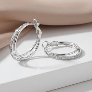 May include: A pair of silver hoop earrings with a double-hoop design. One hoop is adorned with small, clear gemstones. The earrings are presented on a white surface with a neutral-toned fabric in the background.