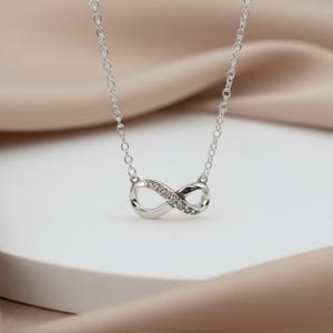 May include: A silver necklace with an infinity symbol pendant. The pendant is embellished with small, clear gemstones. The necklace is presented on a white surface with a soft, neutral-toned fabric in the background.