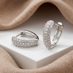 May include: A pair of silver hoop earrings, each encrusted with multiple small, clear gemstones. The earrings are displayed on a white surface with a neutral-toned fabric in the background. The gemstones sparkle, catching the light.