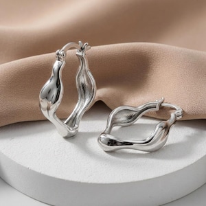 May include: A pair of silver-toned hoop earrings featuring an irregular, organic, and fluid shape with a polished, reflective surface. Each earring is constructed with a hinged post mechanism for secure closure, showcasing a smooth, sculptural design.