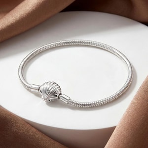 May include: A silver-toned snake chain bracelet featuring a central clasp shaped like a seashell. The bracelet has a smooth, flexible metallic texture and a polished finish, with the shell clasp serving as the primary focal point of the design.