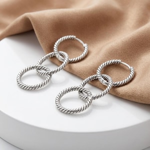 May include: A pair of silver-toned hoop earrings, each featuring a trio of interlocking circular rings. The metal surface of every ring displays a distinct twisted rope texture, creating a continuous, linked design that hangs in a vertical arrangement.