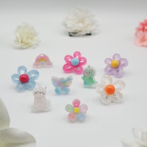 Könnte beinhalten: Eine Sammlung bunter Harzanhänger in verschiedenen Formen, darunter Blumen, eine Katze, ein Schmetterling und ein Regenbogen. Die Anhänger sind in den Farbtönen Rosa, Blau, Gelb und Weiß gehalten und haben ein durchscheinendes Aussehen. Weiße Blumen sind im Hintergrund.