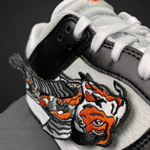 Könnte beinhalten: Nahaufnahme eines weißen, schwarzen und orangefarbenen Sneakers. Der Schuh hat einen detaillierten gestickten Aufnäher eines Tigerkopfes und -körpers. Die Schnürsenkel sind weiß und der Schuh hat eine schwarze Lederausstattung.