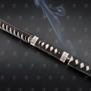 May include: A sword handle featuring a black cord-wrapped grip with white ray skin panels visible underneath. The handle is accented by two silver-colored metal collars with engraved geometric patterns, with thin wisps of smoke rising above.