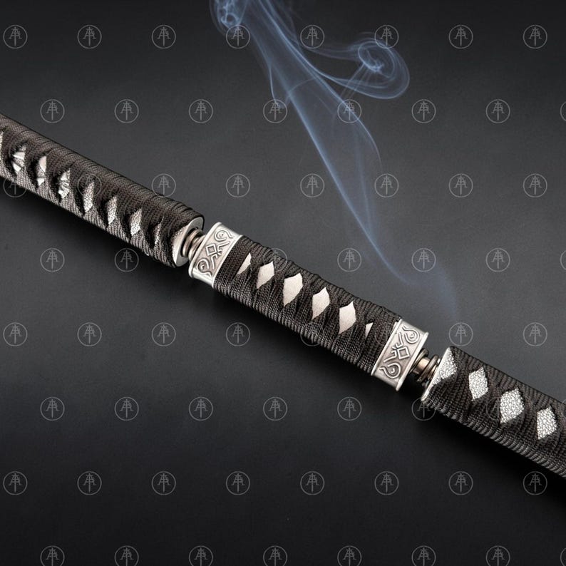 May include: A sword handle featuring a black cord-wrapped grip with white ray skin panels visible underneath. The handle is accented by two silver-colored metal collars with engraved geometric patterns, with thin wisps of smoke rising above.