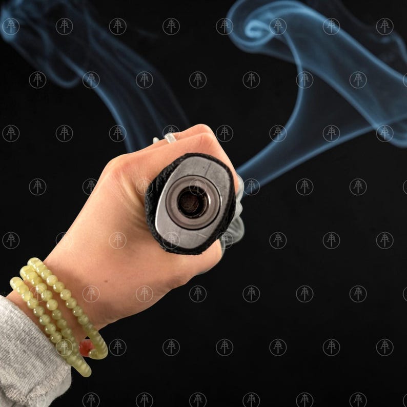 May include: A cylindrical object with a black textured grip and a metallic circular end cap featuring a central opening. Wisps of blue-tinted smoke emanate from the opening against a black background. Three light green beaded bracelets are visible.