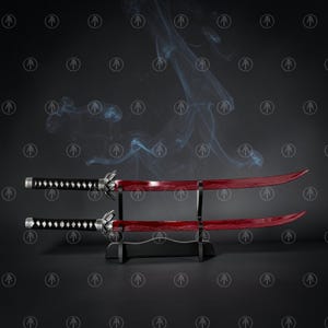 May include: Two katana swords with deep red blades and black handles featuring silver-toned accents. The swords are displayed horizontally on a black tiered stand, with wisps of smoke rising behind them against a dark, patterned background.
