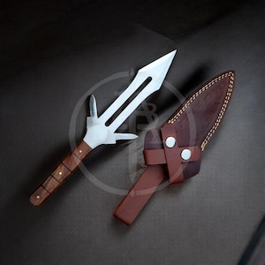 Könnte beinhalten: Ein handgefertigtes Speerspitzenmesser mit Holzgriff und Lederscheide. Das Messer hat eine silberne Klinge mit einem einzigartigen Design. Die Scheide ist braun und hat einen Riemen mit zwei silbernen Knöpfen.