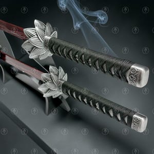May include: Two swords with dark red blades and black cord-wrapped handles. Each handle features a silver-toned metal tsuba shaped like a lotus flower and a silver pommel with engraved patterns. The swords are positioned on a dark display stand.