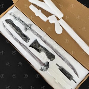 May include: A disassembled sword set packaged in a brown cardboard box with white foam inserts. The set includes a long blade, a handle, and a crossguard, all individually wrapped in clear plastic. The foam is molded to fit each component securely.