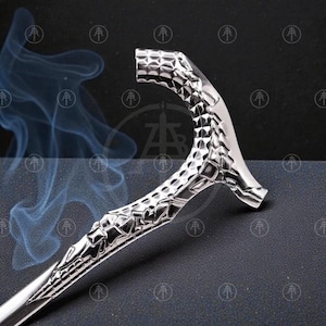 May include: A silver-colored cane with a textured handle and a curved design. The handle has a geometric pattern, and the cane appears to be made of metal. The cane is set against a dark background with a subtle smoke effect.
