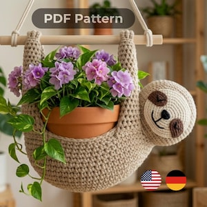 Puede incluir: Un colgador de plantas de perezoso tejido a mano con un tiesto de terracota lleno de flores moradas y follaje verde. El perezoso tiene un cuerpo beige, ojos marrones y una cara sonriente. El texto "PDF Pattern" está en la parte superior.