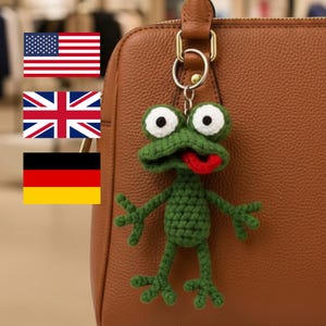 Puede incluir: Un llavero de rana verde tejido a ganchillo, con ojos blancos y una lengua roja. La rana está sujeta a un bolso de mano de cuero marrón. El llavero es un accesorio caprichoso.