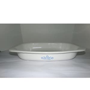 May include: A rectangular white ceramic baking dish with rounded corners and integrated side handles. The exterior features a small, centered blue floral pattern consisting of stylized flowers and leaves on the front side.