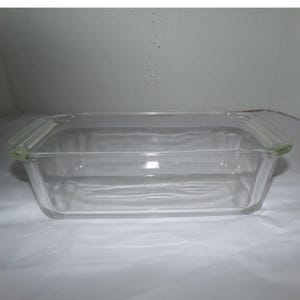 Puede incluir: Un molde rectangular de vidrio transparente con paredes lisas y asas integradas en ambos extremos. El recipiente cuenta con una base plana y bordes ligeramente ensanchados, ofreciendo un diseño sencillo y funcional para hornear.