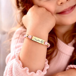 May include: A gold-toned metal identification bracelet featuring a rectangular plate engraved with the name Sophia and a small butterfly icon. The plate is attached to a light pink cord with two small gold-toned spherical beads on each side.