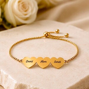 Op de afbeelding: Een verstelbare goudkleurige armband met drie verbonden hartvormige bedels. Elk hart is gegraveerd met een naam in zwarte schrijfletters: David, Mom en Sarah. De armband heeft een fijne ketting en een schuifsluiting met kraal.