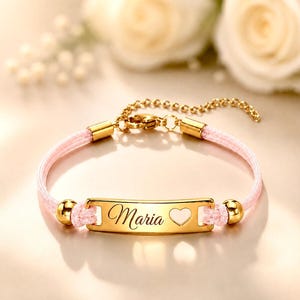 Può includere: Un braccialetto color oro con un cordino rosa chiaro e una piastra metallica rettangolare centrale incisa con il nome Maria e un piccolo cuore ritagliato. Il braccialetto include perline color oro, una chiusura a moschettone e una catenella.