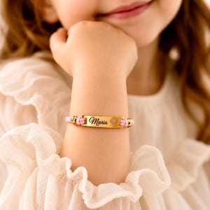 Pode incluir: Uma pulseira de identificação de metal dourado com o nome Maria em letras cursivas pretas e um pequeno recorte em forma de coração. A placa retangular central está ligada a um cordão rosa claro com contas douradas nas laterais.