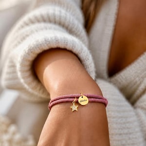 Könnte beinhalten: Ein doppeltes Armband aus malvenfarbener Kordel mit zwei goldfarbenen Anhängern: einer runden Scheibe mit der Gravur Amelia und einem kleinen sternförmigen Anhänger, der mit klaren Steinen besetzt ist, befestigt mit einem goldfarbenen Verschluss.