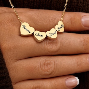 Puede incluir: Un collar de tono dorado con cuatro pequeños dijes en forma de corazón suspendidos de una cadena fina. Cada corazón está grabado con un nombre en letra cursiva negra: Grandma, Claire, David y Emily. Los dijes están dispuestos en fila.
