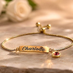 Könnte beinhalten: Ein verstellbares Armband mit goldfarbener Boxkette und einem zentralen rechteckigen Steg, der mit dem Namen Charlotte in Schreibschrift graviert ist, ergänzt durch einen kleinen, runden, rosafarbenen Edelsteinanhänger an der Seite.