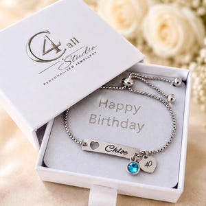 Op de afbeelding: Een zilverkleurige verstelbare kettingarmband met een rechthoekige balk gegraveerd met de naam Chloe en een hartvormige uitsparing. De armband heeft een kleine blauwe kristallen bedel en een hartvormige bedel met het getal 40, gepresenteerd in een witte doos.