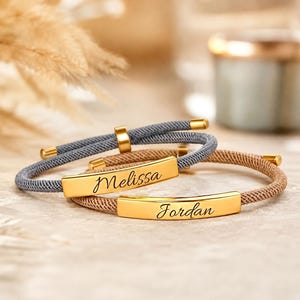 Puede incluir: Dos pulseras de cordón ajustable con barras de metal dorado. Una pulsera tiene un cordón gris con el nombre Melissa grabado en letra cursiva negra, y la otra un cordón beige con el nombre Jordan grabado en letra negra sobre la barra.