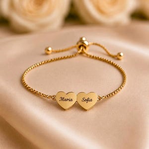 Könnte beinhalten: Ein verstellbares goldfarbenes Armband mit einer feinen Boxkette und zwei herzförmigen Anhängern. Auf jedem Herz ist ein Name in schwarzer Schreibschrift eingraviert, links Maria und rechts Sofia. Das Armband verfügt über einen Schiebeverschluss.