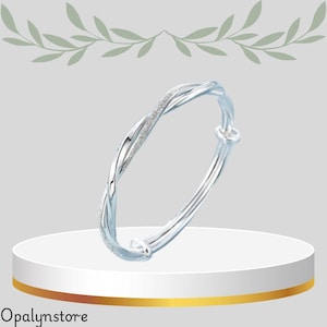 May include: A silver bangle bracelet with a twisted design. One side of the bracelet has a textured, glittery appearance. The bracelet is displayed on a white pedestal with a gold band.