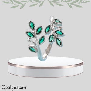Puede incluir: Anillo plateado con diseño de hojas y gemas verde esmeralda. El anillo tiene un diseño delicado y abierto, con hojas que envuelven el dedo. Se exhibe sobre un pedestal blanco con un borde dorado. Joyería.