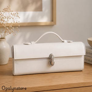May include: A rectangular off-white handbag with a smooth finish, featuring a single top handle and a central silver-toned metal clasp closure. The bag has a structured horizontal design with clean stitched edges and a secure flap front.