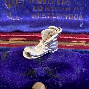 Può includere: Un piccolo ciondolo a forma di stivale in argento, esposto all'interno di un portagioie vintage. Il ciondolo è dettagliato. La scatola ha un interno in velluto blu e il testo "BOWMAN LTD WATCHMAKERS JEWELLERS LONDON W. WEST YOO" è visibile.