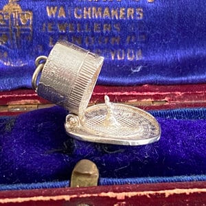 Op de afbeelding: Een zilveren bedel in de vorm van een emmer en een bord, gepresenteerd in een open vintage rode sieradendoos met een blauwe fluwelen binnenkant. De doos toont de fijne details van de bedel en de reliëftekst "BOWMAN LTD WATCHMAKERS JEWELLERS".