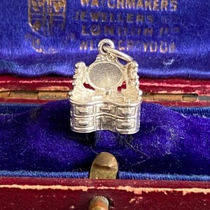May include: A silver charm depicting a dressing table with a mirror and drawers, displayed on blue velvet within a red jewellery box. The charm is detailed and likely vintage.