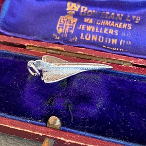 May include: A silver-toned metal charm shaped like a space shuttle, featuring a small jump ring at the rear. The charm is resting inside a rectangular, hinged jewelry box with a deep red exterior and a vibrant blue velvet interior lining.