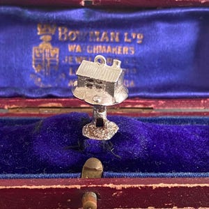 Puede incluir: Un charm de plata con forma de casa, presentado en un joyero vintage rojo y azul. El charm tiene detalles de ventanas y una chimenea. La caja tiene letras doradas que dicen "Bowman Ltd Watchmakers".