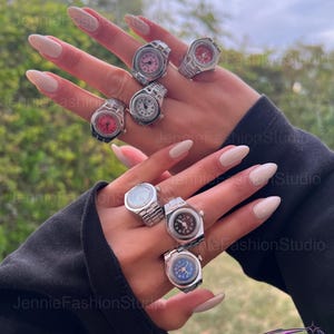 May include: A selection of silver watch rings with faces in shades of pink, red, white, and blue. The rings are shown on a hand with long, polished nails. The background is out of focus.