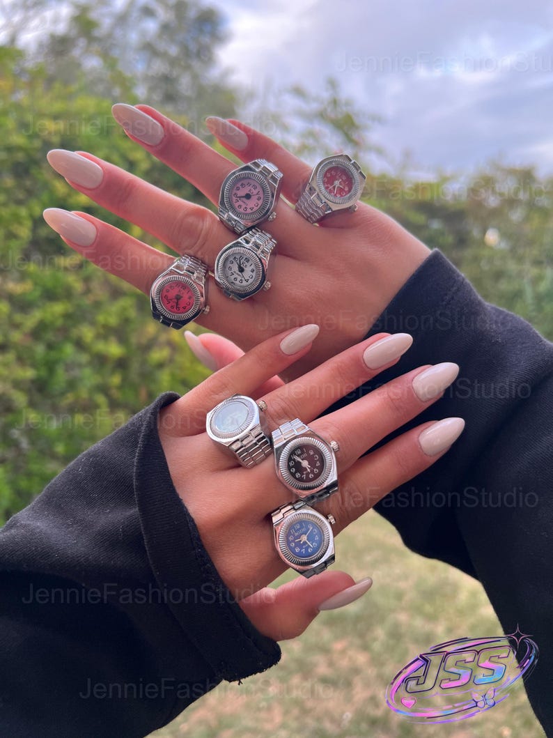 May include: A selection of silver watch rings with faces in shades of pink, red, white, and blue. The rings are shown on a hand with long, polished nails. The background is out of focus.