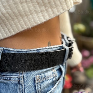 May include: Black leather belt with embossed floral pattern, worn with blue jeans. The belt has a silver buckle and is a stylish accessory.
