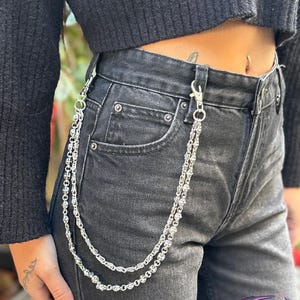 May include: Silver wallet chain with a decorative design, attached to black denim jeans. The chain is made of multiple linked segments and has a lobster clasp. The jeans are a dark wash and have visible pockets and a button closure.