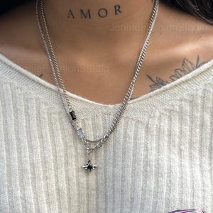Puede incluir: Un collar plateado de varias capas con un pequeño colgante. El collar tiene un colgante de piedra negra y una cadena con piedras claras y negras rectangulares y cuadradas. La palabra "AMOR" está tatuada en el cuello.